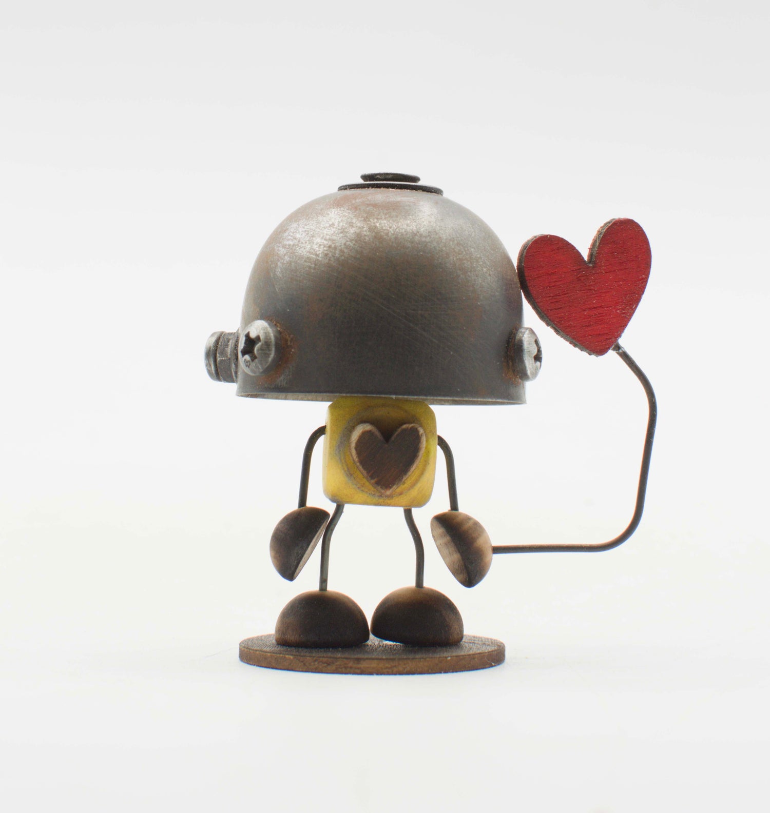 Una figurina robot minibot metallica con testa a cupola argento arrugginita torso giallo con cuore ritagliato e arti in legno che tiene un cuore rosso su filo