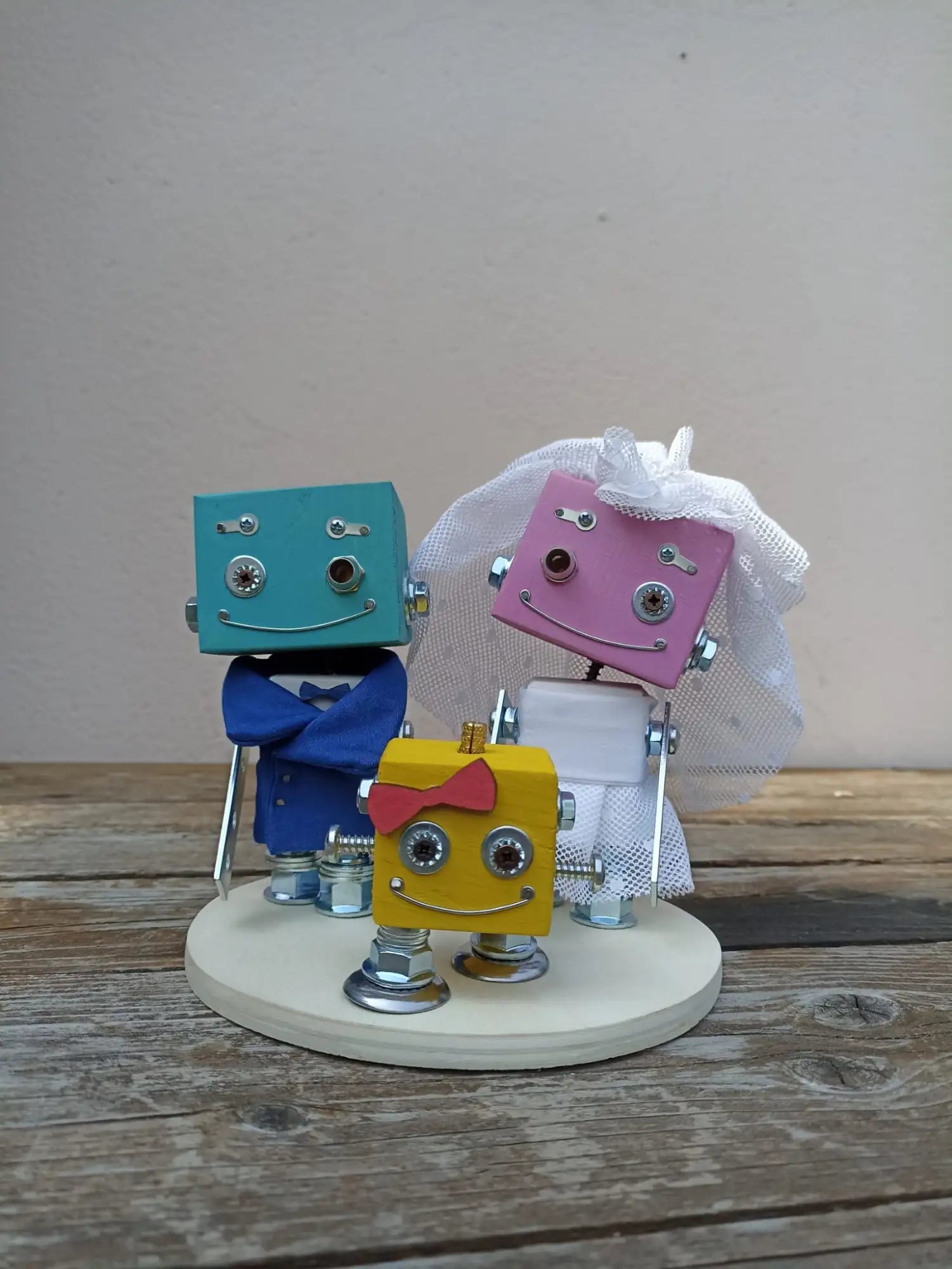 Un affascinante trio di colorati robot come decorazioni per torte nuziali, con uno sposo turchese in abbigliamento blu, una sposa rosa con velo in rete bianca e papillon, e un bambino giallo con un fiocco rosso.
