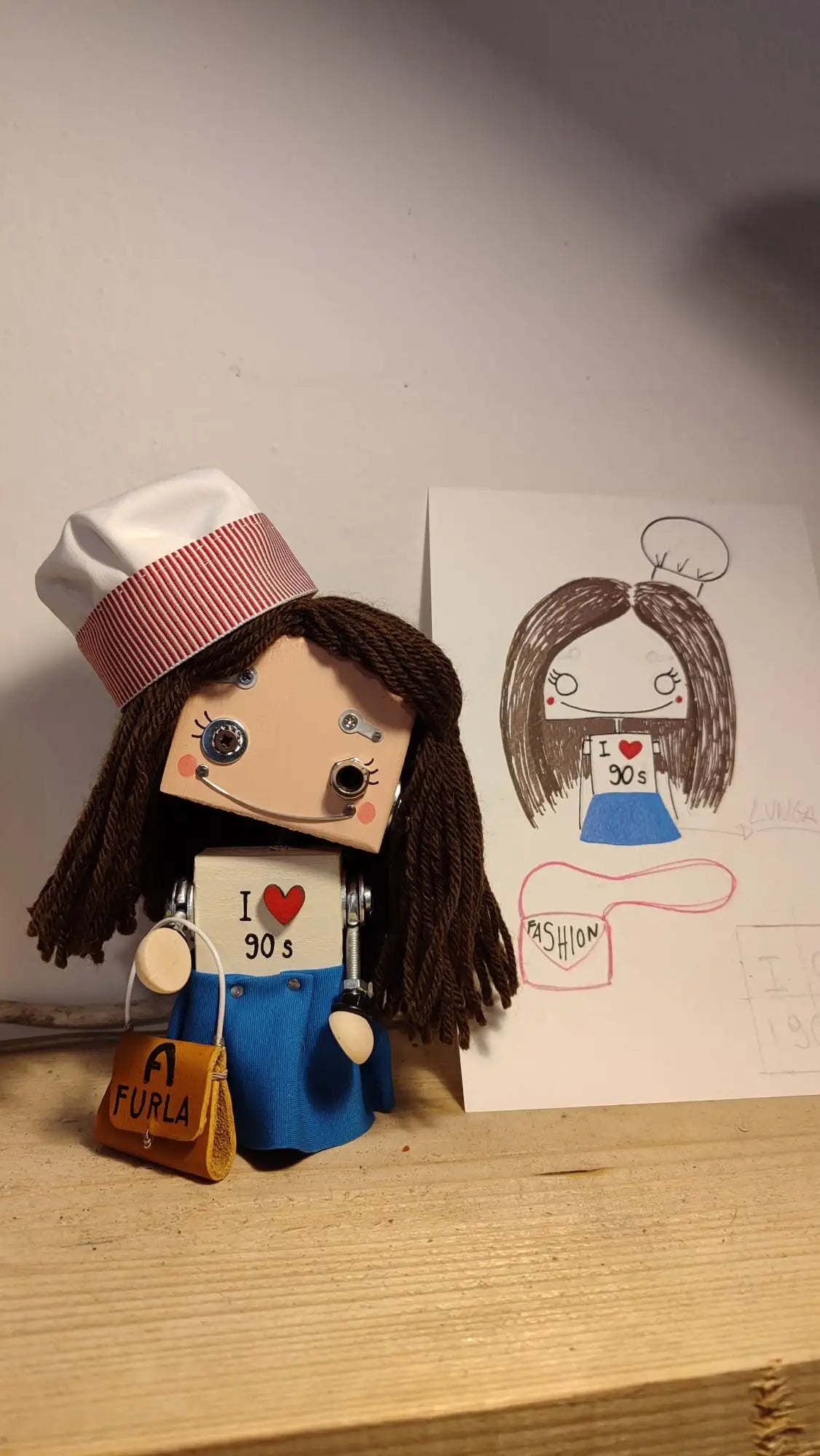 Una bambola fatta a mano con una testa quadrata, capelli di lana marrone e un cappello da chef bianco con strisce rosse, che indossa una tuta blu e tiene in mano una borsa Furla arancione.