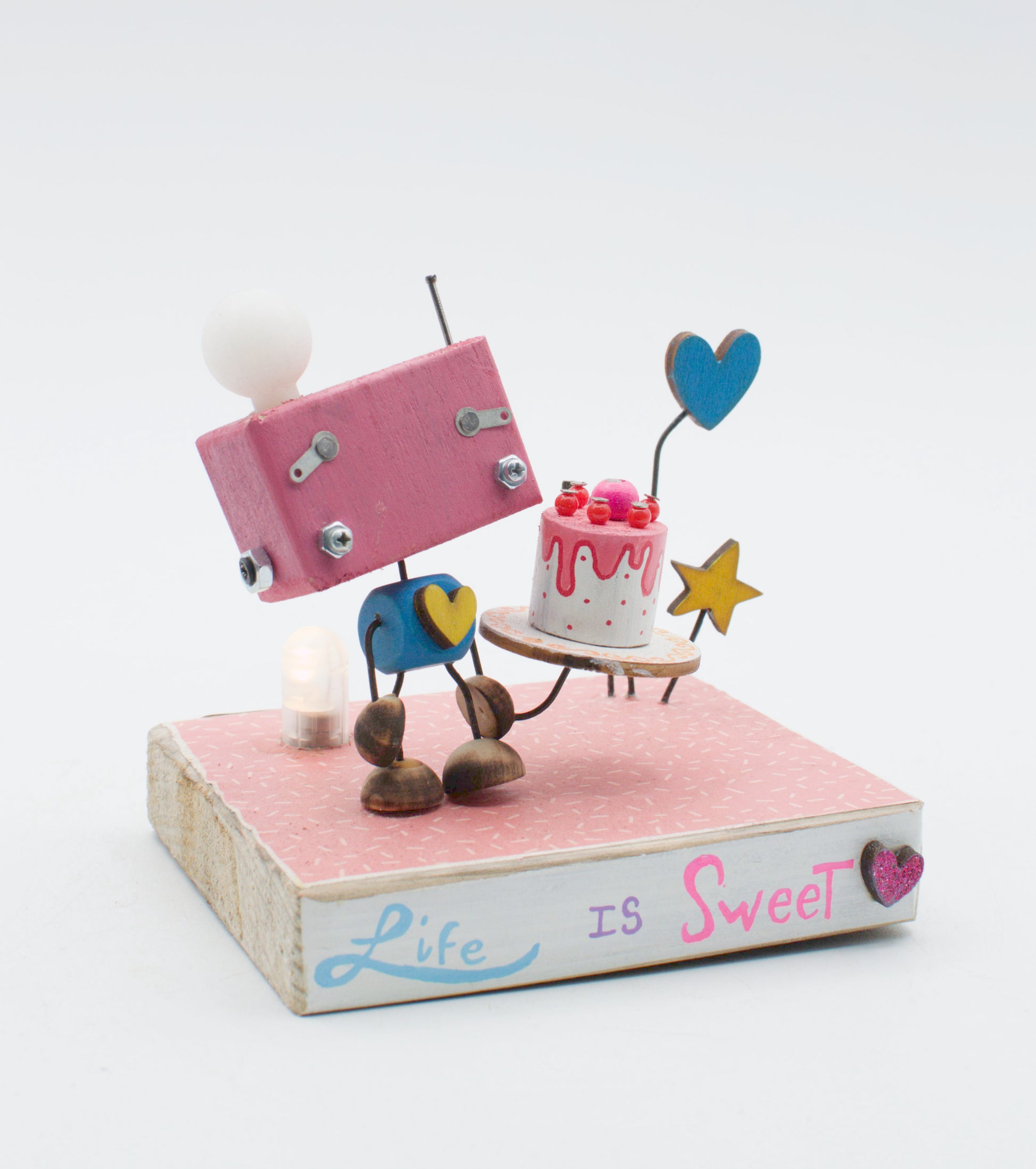 Diorama scultura luminosa stile robot fatto a mano rosa con corpo blu e piedi marroni che tiene una torta su vassoio di legno