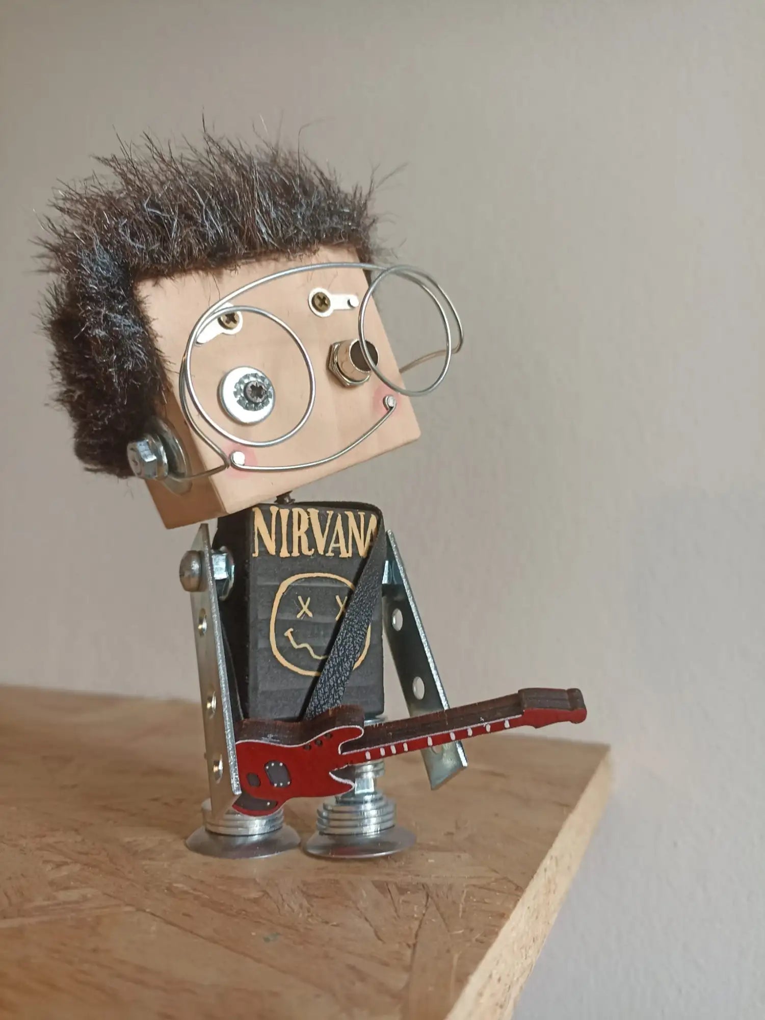 Una figurina di robot metallico fatta a mano con testa quadrata, capelli marroni di pelliccia sintetica, occhiali con montatura di filo metallico e maglietta della band Nirvana, che tiene in mano una chitarra elettrica rossa.