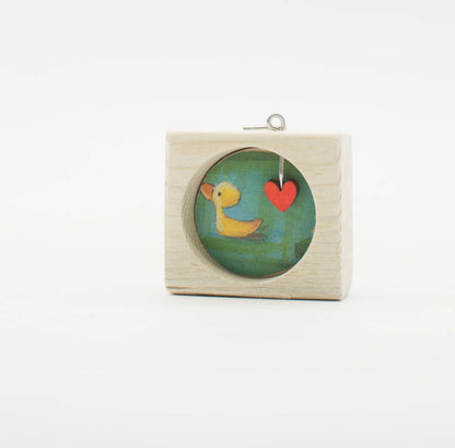 Piccolo quadro artigianale in legno chiaro con finestra tonda che mostra un’anatra gialla dipinta e un cuore rosso su sfondo verde