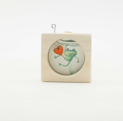 Piccolo quadro artigianale in legno con inserto di vetro circolare raffigurante una rana verde che tiene un cuore arancione