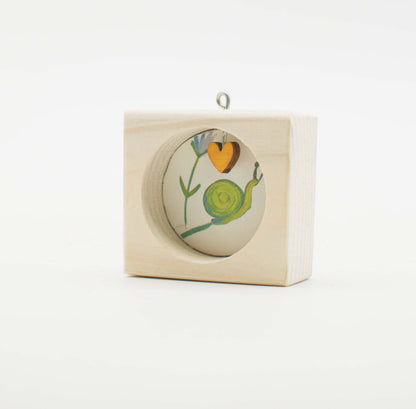 Piccolo quadro artigianale in legno con lumaca verde e cuore arancione su sfondo bianco con appendino argento