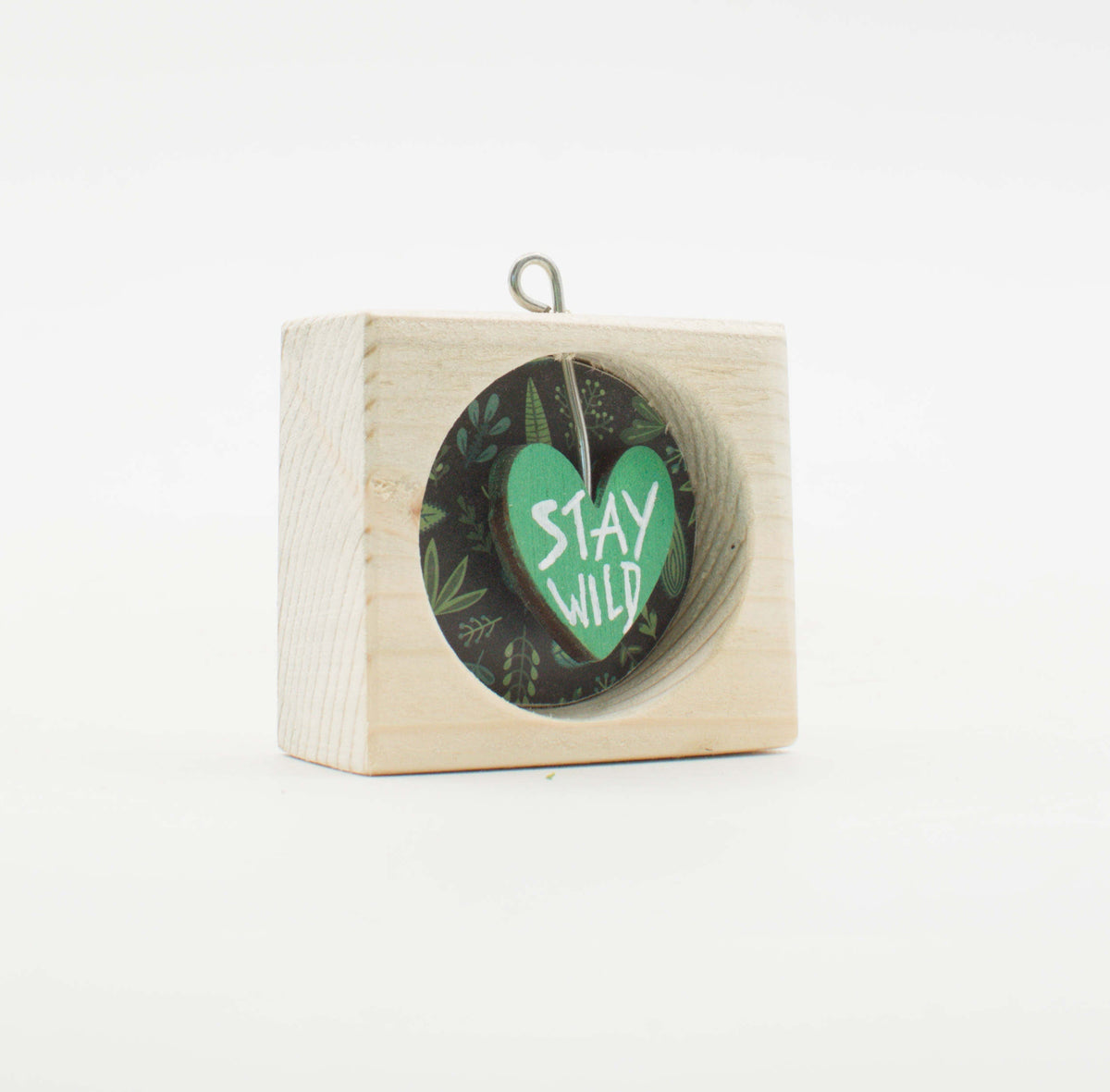 Un piccolo quadro artigianale in legno con finestra circolare verde cuore stay wild su sfondo fogliame scuro