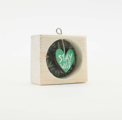 Un piccolo quadro artigianale in legno con finestra circolare verde cuore stay wild su sfondo fogliame scuro
